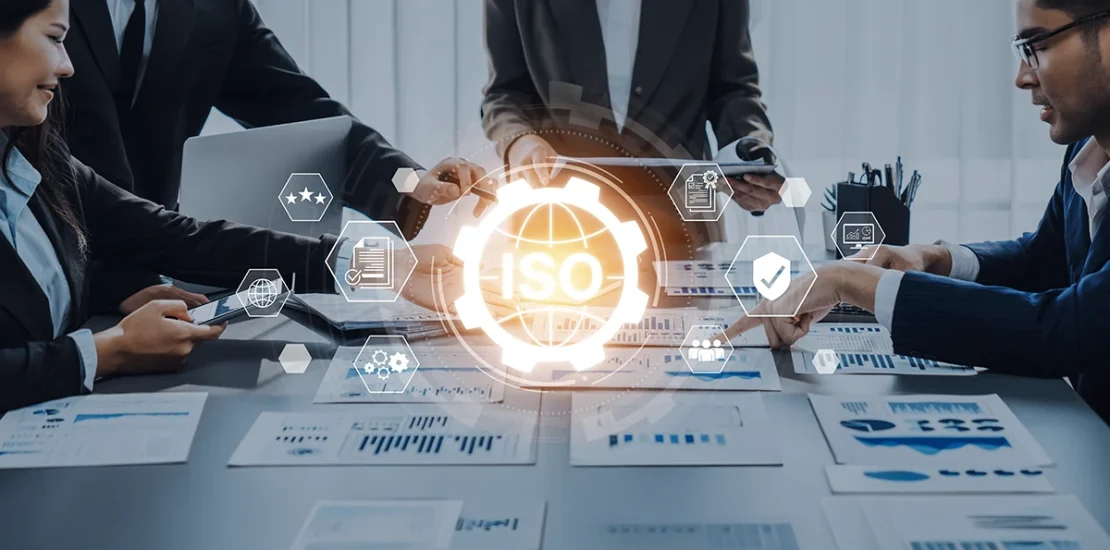 A diverse team of professionals in a UAE office collaborating on digital documents. A central "ISO" digital overlay with a gear and globe icon illustrates the implementation of Integrated Management Systems (IMS) in UAE.