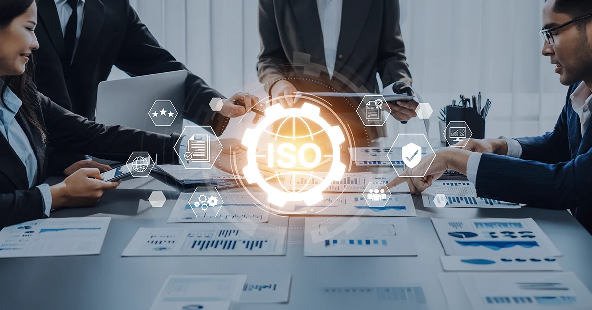 A diverse team of professionals in a UAE office collaborating on digital documents. A central "ISO" digital overlay with a gear and globe icon illustrates the implementation of Integrated Management Systems (IMS) in UAE.