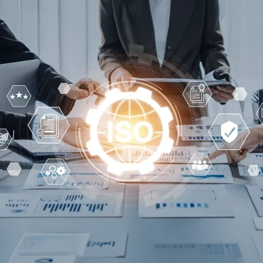A diverse team of professionals in a UAE office collaborating on digital documents. A central "ISO" digital overlay with a gear and globe icon illustrates the implementation of Integrated Management Systems (IMS) in UAE.